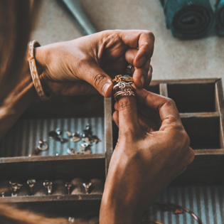 A set of hands over a jewellery box filled with rings that are holding three rings on one thumb
